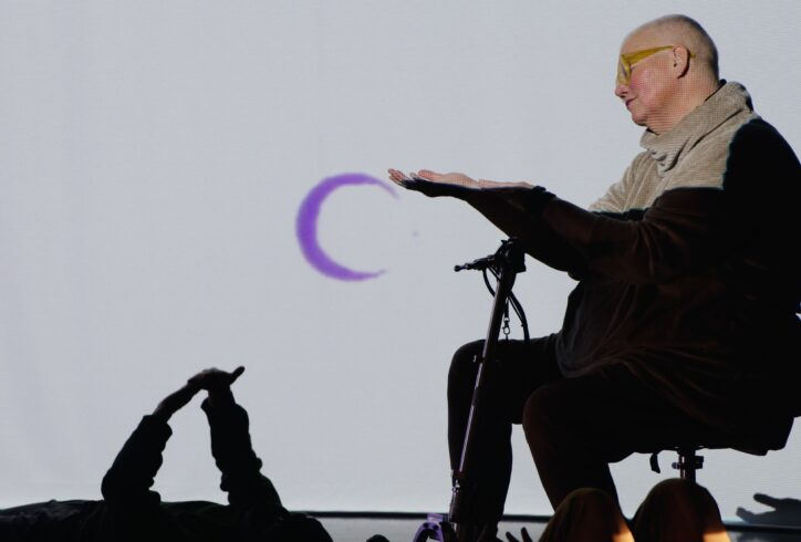 Petra, a white queer disabled cis woman of size sits on her scooter, and reaches out her hand to a video projection of a purple moon. Below, a floor dancer reaches up and touches their own hands. Photo courtesy of the artist.