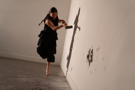 Martita Abril takes a sledgehammer to a wall while balancing on one foot. She wears a black dress and her hair in a braid. Photo by Julieta Cervantes.