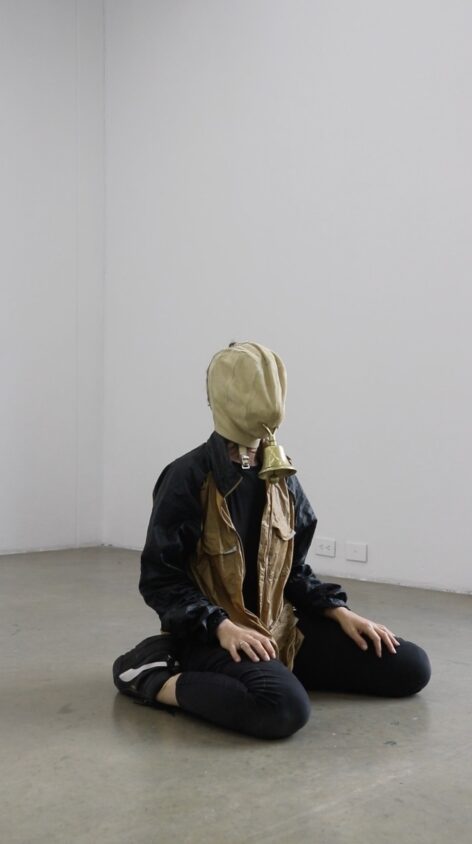 A body sits on the floor, legs bent backwards, hands resting
onto of its knees. The body is wearing black puma trainers,
black trousers, a black and gold jacket. The body wears a
mask made of suede over its face, where the mouth would
be, hangs a copper bell. The space behind the body is
painted white and the floor is painted grey. Photo courtesy of the artists.