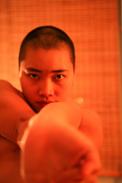 Portrait of Shannon Yu. Shaved head asian with hands reach towards camera. Red light washes over Shannon. Photo by Shannon Yu.