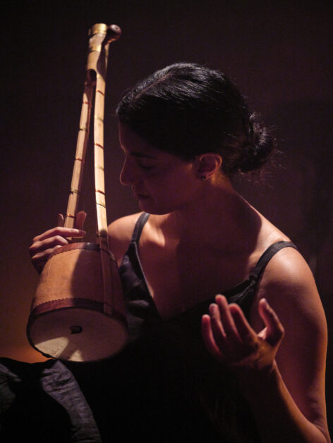 Samita, an Indian-American woman, performs seated, wearing a black dress and her hair pulled back in a bun. She plays an ektaara, a one-stringed instrument, and her eyes are closed in a posture of listening. Photo courtesy of Rachel Topham Photography.