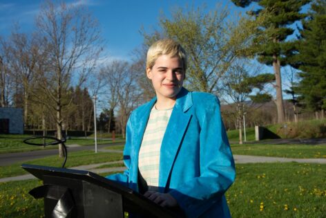 Kari, a white nonbinary person with short bleached hair, stands at a
podium smiling with a teal suede blazer on. In the background, blue skies, trees and grass.