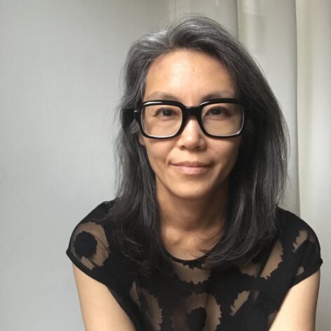Kay Takeda looks into the camera. Her hair is varying shades of gray and she wears black rectangle glasses and a sheer black top. She has a soft smile. Photo courtesy of Kay Takeda. 