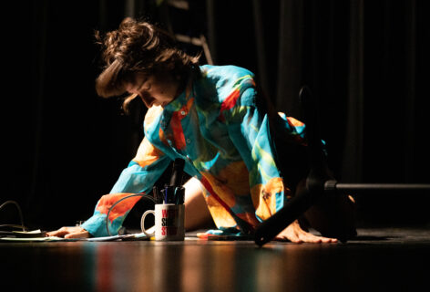 Tatyana wears a bright patterned shirt with arms pressed firmly into the floor. Her eyes are closed and her open mouth hovers over a microphone. Blurred hair suggests movement. Photo by Maria Baranova.