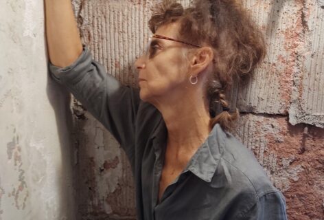 Photo of Vicky leaning on an exposed cinder block wall. She is in profile with one arm resting on the wall in front of her. She is wearing a blue button-down top. Photo courtesy of the artist.