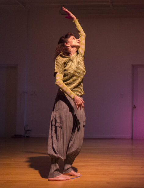 Cathy Weiss performs. She is in movement, standing with one arm reaching upwards and head leaning diagonally. She wears gray baggy pants and a green top. Photo by A Hitzenberger.