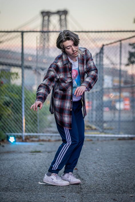 evan dances slowly in a park on the Lower East Side, with the Williamsburg Bridge in the background. evan has medium-length brown and gray hair and wears a plaid wool shirt over a sweatshirt depicting people bathing in the top of Mt. Fuji. Photo by Shintaro Ueyama.