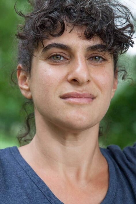 Portrait of Rebecca Pappas. She looks straight into the camera with a neutral expression. She has hazel eyes, dark curly hair with bangs and wears a dark blue top. Photo courtesy of the artist.
