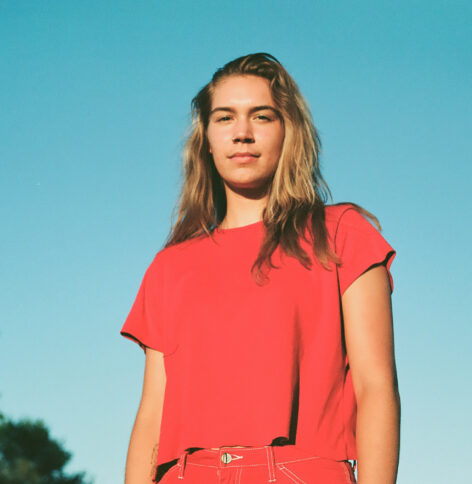 Maia, a white woman, wears a red outfit. She is against a clear sky, looking directly into the camera. Photo courtesy of the artist.