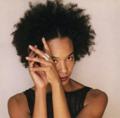 Edisa poses for a photo. Her hands are held up covering one side of her face. Her eyes look into the camera. Her hair is in an afro and she wears a dark sleeveless top. Photo courtesy of Edisa Weeks.