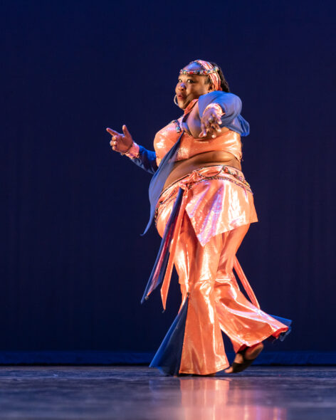 A full-figured Black woman is seen on stage from stage left. She is looking left from the side at the camera with one arm extended toward it, the other pointing toward the audience. She wears a metallic orange and royal blue costume. The pants are bell bottoms with an orange skirt overlay and blue sheer accents. The top is a halter-bra with multi-color beading outlining the collar. There is a sheer blue belly drape in front. Her orange headband also includes the multi-color beading