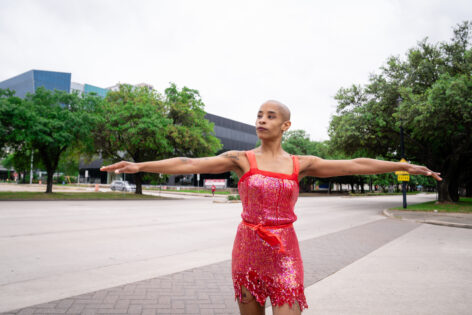 Jasmine, a Black person with a shaved head stands at the intersection of Main Street and Portland, wearing a red sequins dress. Their arms are outstretched to their sides. There are trees in background with lush forest green leaves and grayish bark trunks. Photo by Jakayla Monay