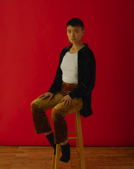 Cherrie has short black hair, wearing a black shirt, with white shirt inside, yellow pants, sitting on a stool in front of a red backdrop. Photo by Jason Lê