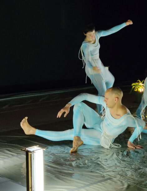 Photo from a performance. Dancers Demetris Charalambous & Kalliope Piersol dance wearing light blue unitards. Demetris sits with one leg extended out and the other crossed the first. Kalliope is on mid movement seemingly falling side ways with their arms outstretched. Photo by Jeremy Nelson.