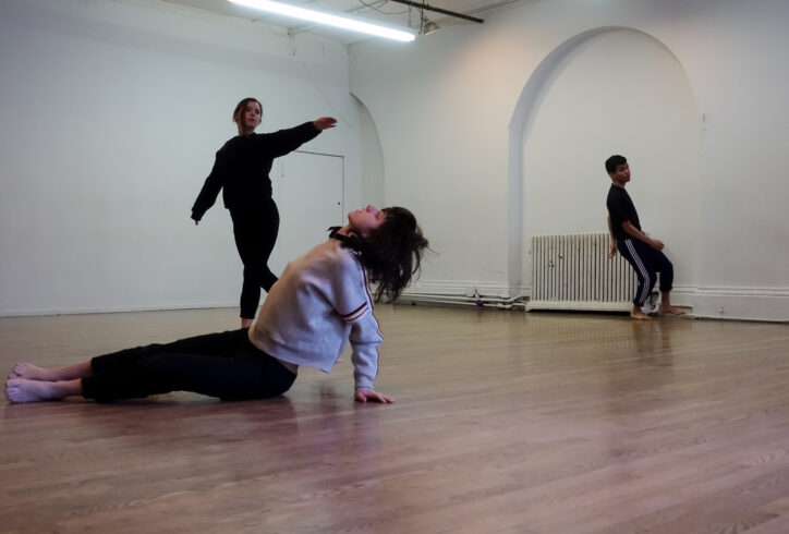 Three dancers move in the space. In the foreground a dancer in a gray sweatshirt and dark pants sits with their head thrown back and arms supporting their torso. In the background two other dancers in black move around. One extends their arm away from their body. Harry Shunyao Zhang.