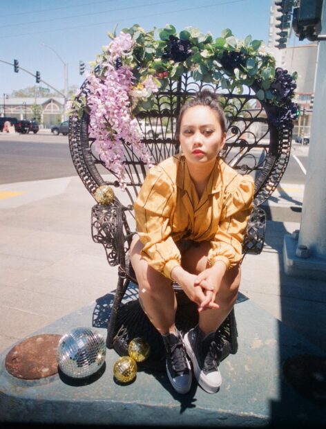 Portrait of Kmay. They sit on a peacock chair decorated with flowers. The chair is placed on a busy corner of their childhood neighborhood. They lean forward in a gold blouse with small disco balls surrounding them. Photo by Tunuviel Love captured on film.