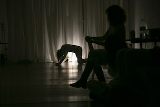 Silhouette of dancer on hands and knees in front of a white curtain, a silhouette of a seated person in the foreground