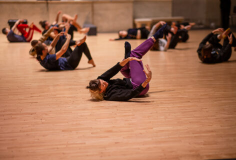 A class photo at the Judson Memorial Church. In the foreground a dancer lays on their back in a contraction spiraling their torso while holding their arms with their arms curved fingers nearly touching. Their legs are crossed and their feet are pointed. The dancer wears purple sweats and a black long sleeve top. In the background several other dancers are doing the same pose. Photo by Rachel Keane.