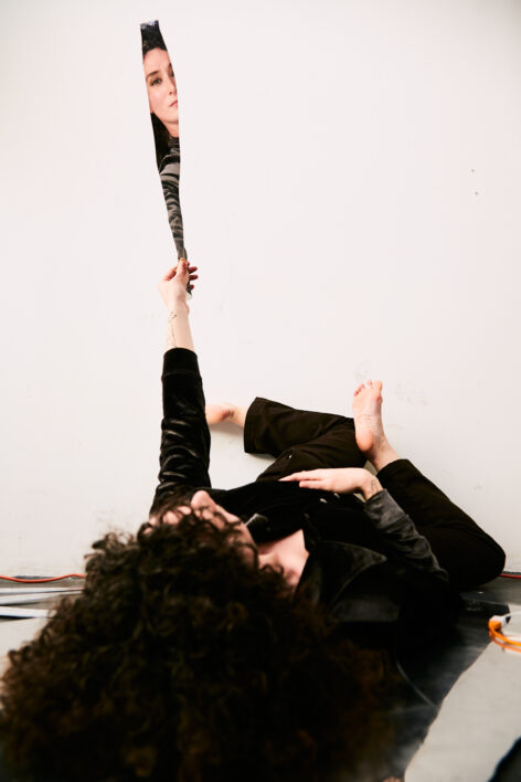 Photo by Whitney Browne. A white woman dressed in dark shirt and pants lies on ground in room with white walls and floor; woman's left arm is outstretched, lifted up holding a piece of glass. In the reflection you can see her face.