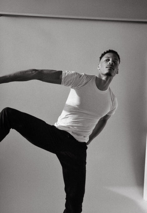  A black and white photo of Jelani Taylor. He wears a white tshirt and black pants. His  right leg and arm extend away from his body. Photo by Emily Farthing.