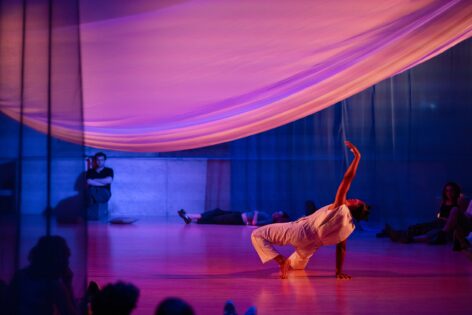 Brown skinned dancer in white sleeveless jump suit in a back bend with hands extended toward white suspended fabric in a rounded shape, Lighting is a pinkish purplish hue and back drop is the Judson Church dance space. Photo by Emily Farthing
