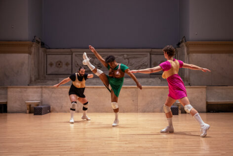 Photo from Blaze Ferrer's performance for Movement Research at the Judson Church. Three dancers wearing prosthetic breasts over dresses move in the space swinging their limbs in different directions. Photo by Rachel Keane