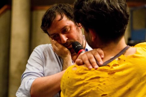 Levi, a light-skinned, dark-haired male is looking down and to his left, while his hand is placed on the shoulder of another dark-haired male whose back is to the viewer. This figure holds a microphone to Levi's mouth and has his hand on Levi's right cheek. Both are standing before a blurred grey background. Photo by Alex Escalante