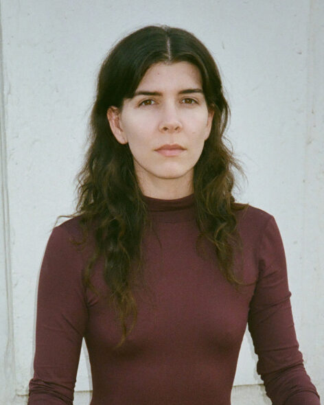 The image shows a woman with long, dark, wavy hair that falls past her shoulders. She is wearing a fitted, long-sleeved, maroon turtleneck shirt. Her facial expression is neutral, with a calm and composed demeanor. Her skin tone is light, and she has dark eyebrows that frame her eyes. The background is a simple off-white or light-colored wall. Photo by Carolyne Loreé Teston.