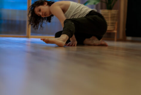 Allie is kneeling on the floor, left leg bent and extended toward the camera. Her toes are flexed off the floor and she is barefoot wearing a white top and green pants. Allie is in the background looking past her foot into the foreground of a light wood floor. Photo courtesy of the artist.