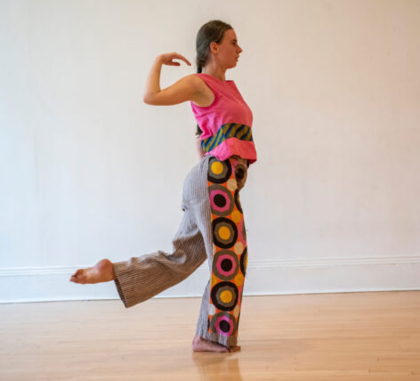 Amanda Kmett'Pendry in movement. She stands on one leg kicking the other behind her with a pointed foot. Her arm is behind her bending at the elbow fingers nearly touching her shoulder. She wears a pink tank top with a printed band. Her pants are black and white stripped with pink yellow and black patterned circles on the side Photo by Amanda Kmett'Pendry.