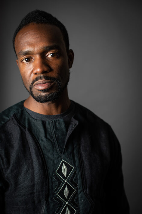 Image of a dark-skinned Black man with trimmed black hair and goatee wearing a black top with white fabric embroidered in cowrie shell design standing in front a slate gray backdrop. Photo by Tara Lynn Pixley.