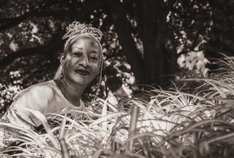 A black and white photo of a black woman with braids close to greenery smiling. Cynthia Oliver photographed by Laura Bianchi for Bogliasco.