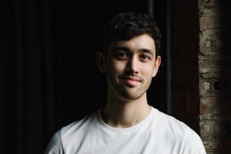 A mixed race, light skin male with short dark wavy hair smiles for a headshot with light cast over the left side of his face. Photo by Da Ping Luo.