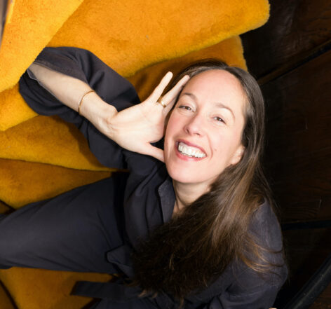 Biba Bell poses on yellow stairs. They smile with their hand to one side of her face.