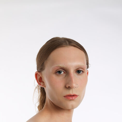 A portrait photo of Buffy looking directly into the camera in front of a white background. Her hair is parted down the middle and pulled back. She is wearing light neutral makeup and a nose ring. Photo by MTHR TRSA @mthrtrsa mthrtrsa.com