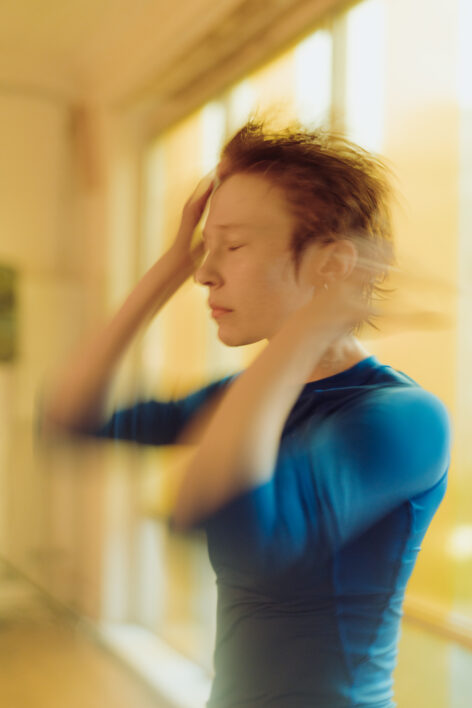 A white person with short red hair closes their eyes while swiping hair away from their face. The edges of the photo blur in motion. Photo by Jenna Maslechko.
