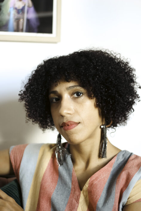 A light brown skin woman with tight bob-length curls and short bangs looks sideways at the camera, wearing long silver tassel earrings and a pastel striped top. Behind her a white wall and a glimpse of a framed photo. Photo credit zakkiyyah najeebah dumas o'neal.