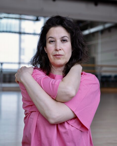 A white woman with brown hair and a pink tshirt hugs her self while standing inside a dance studio. Photo by Amelia Gold