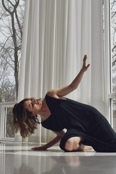Frances rests on the floor, leaning her weight onto her right arm while looking towards her left hand, positioned above her face.  She is wearing a long black dress and her hair is medium-length with Blonde highlights. The room is white and there are barren trees outside the window. Photo by Lee Thomas.