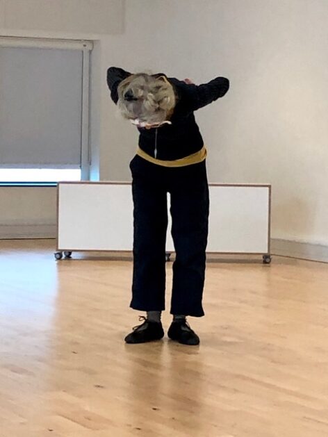 Deborah Hay in black dance clothes and black shoes. She stands on a shiny wooden floor, facing the camera while standing and bent forward so you cannot see her face. Photo courtesy of the the artist.