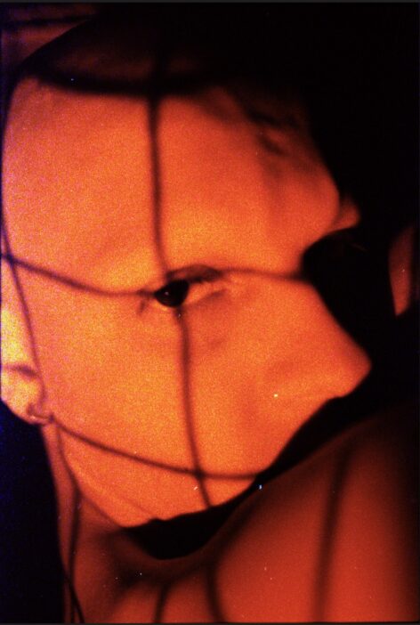 A close up shot of Demetris Charalambous. They are bathed in red light while thin black lines of shadow cross over their face. They look straight into the camera. Photo by Hudson Bohr.