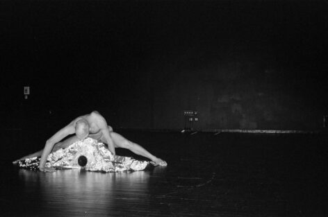 Demetris Charalambous performing. They support their torso on their arms hovering above a metallic mound while their legs are spread at either side of it. Photo courtesy of the artist.  