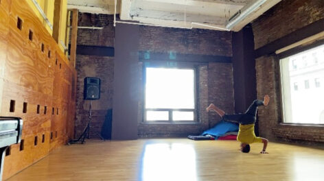A photo of Erick Montes in a brightly lit studio with exposed brick walls. He is in a head stand supporting his weight with his hands pressed into the floor. His legs are spread, knees bent for balance. He wears a yellow shirt and dark pants. Photo courtesy of the artist.