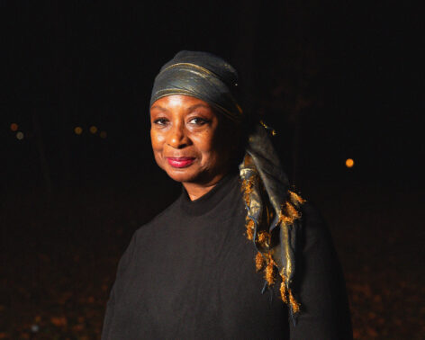 Eva Yaa Asantewaa looking into the camera grinning. She wears a dark gray headscarf with a gold fringe. Photo by Golden for the 2024 Queer|Art Community Portrait Project, assisted by Río Sofia & Andrius Alvarez-Backus
