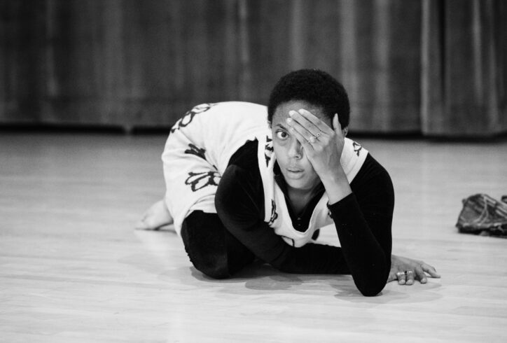 A black and white image of Fana Fraser, a brown-skinned Trinbagonian person, crouched on a hardwood floor with a dark curtain in the background. Fana has her right hand on the floor and her left hand covering one eye. Her right hand wears a turquoise ring on her ring finger and a lapis lazuli ring on her middle finger. Her left hand wears a turquoise ring on her ring finger and a sterling silver ring with two hearts on her middle finger. A crumpled pair of pants sits in the background. Photo by Tony Turner/BAM.