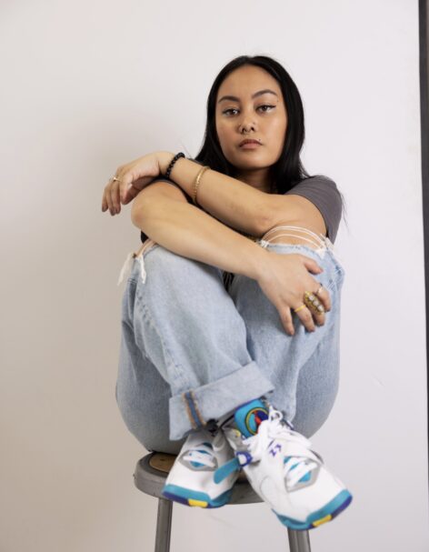 Portrait of Kmay. They sit with their knees pulled to their chest and ankles crossed. They have dark black hair and wear a gray t-shirt, light wash blue jeans and white and blue sneakers. Photo by David Spektor.