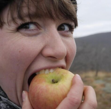 Natalie, looking back over her right shoulder, takes a large bite into a big crisp apple. There is a mountain in the distance. Photo by Fivel Rothberg.