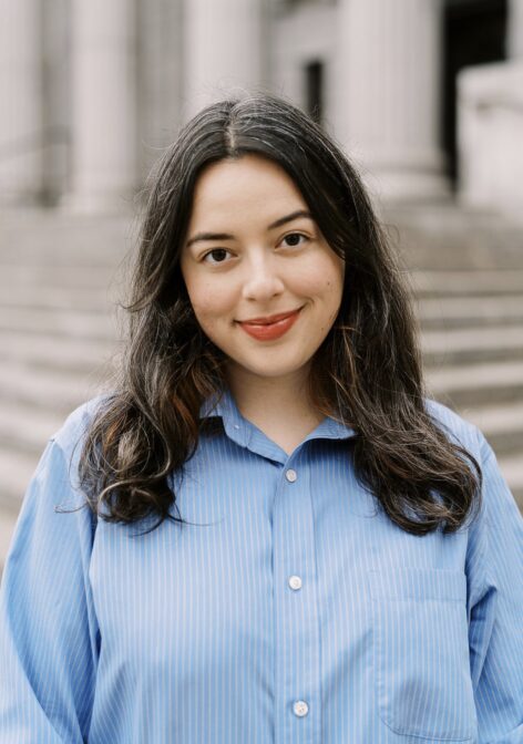 Photo of Maya, who has white skin, dark eyes, and dark hair. She wears a blue button-up shirt with pinstripes and smiles softly, looking into the camera. Photo by Nicole Baas.