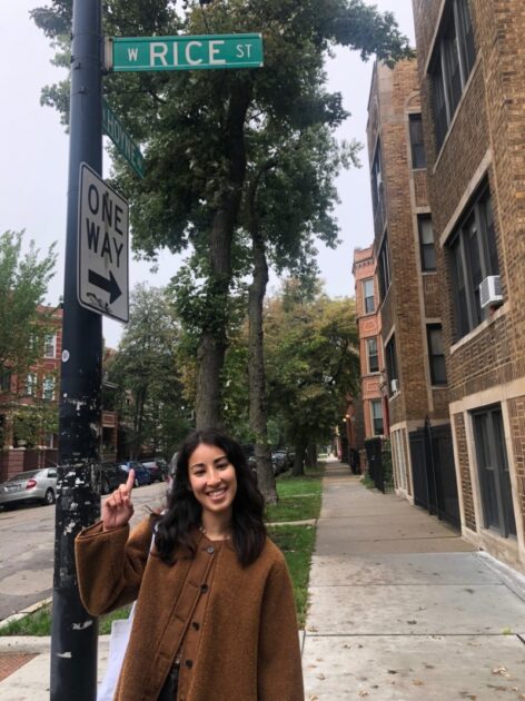 Ayano stands under a street sign that says Rice street in a brown coat. They smile at the camera and point up at the sign. In the background we see a residential street and trees.