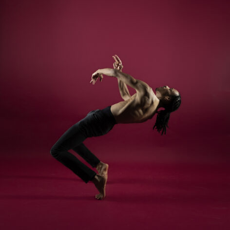 Studio portrait of Jelani Taylor dancing in front of a deep red background. He is on his toes in hinge position with his knees reaching forwards and his torso reaching up and diagonally in counterbalance. His arms wave in front of him. Photo courtesy of the artist.
